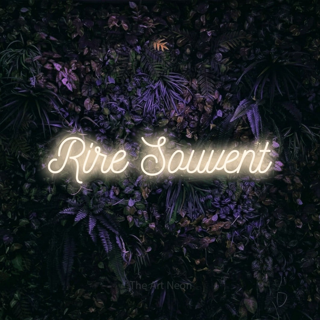 “Rire souvent” LED neon sign in warm white, with ornate cursive lettering, shown against a dark wall with purple flowers.