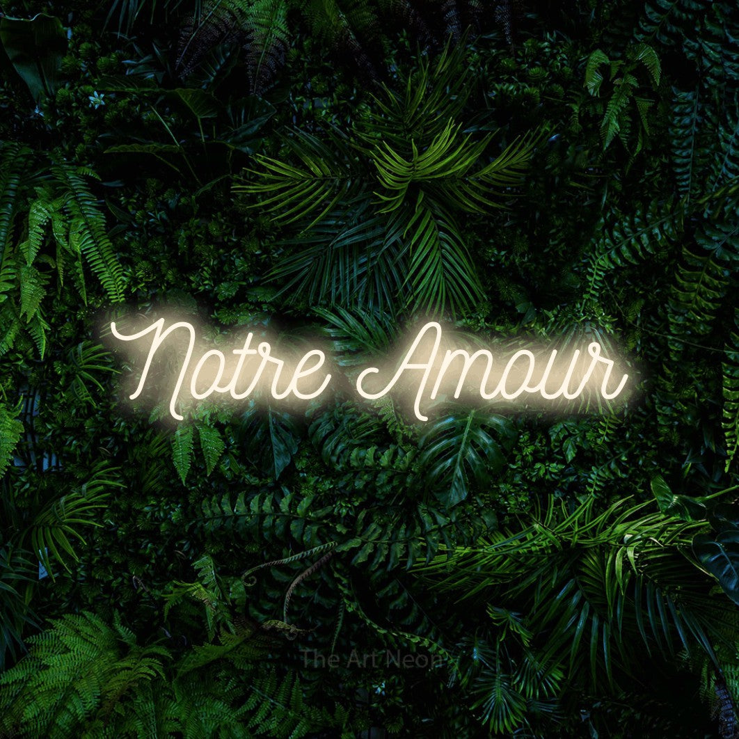 “Notre amour” LED neon sign in warm white, with ornate cursive lettering, shown against dense tropical foliage.