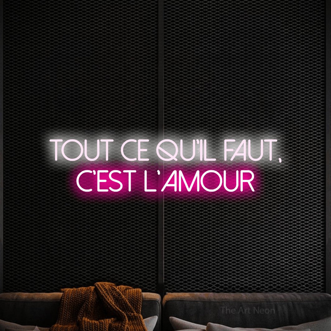“Tout ce qu'il faut, c'est l'amour” LED neon sign in cool white on the upper line and hot pink on the lower line, with thin sans-serif lettering across 2 lines, shown against a modern dark-toned seating area.