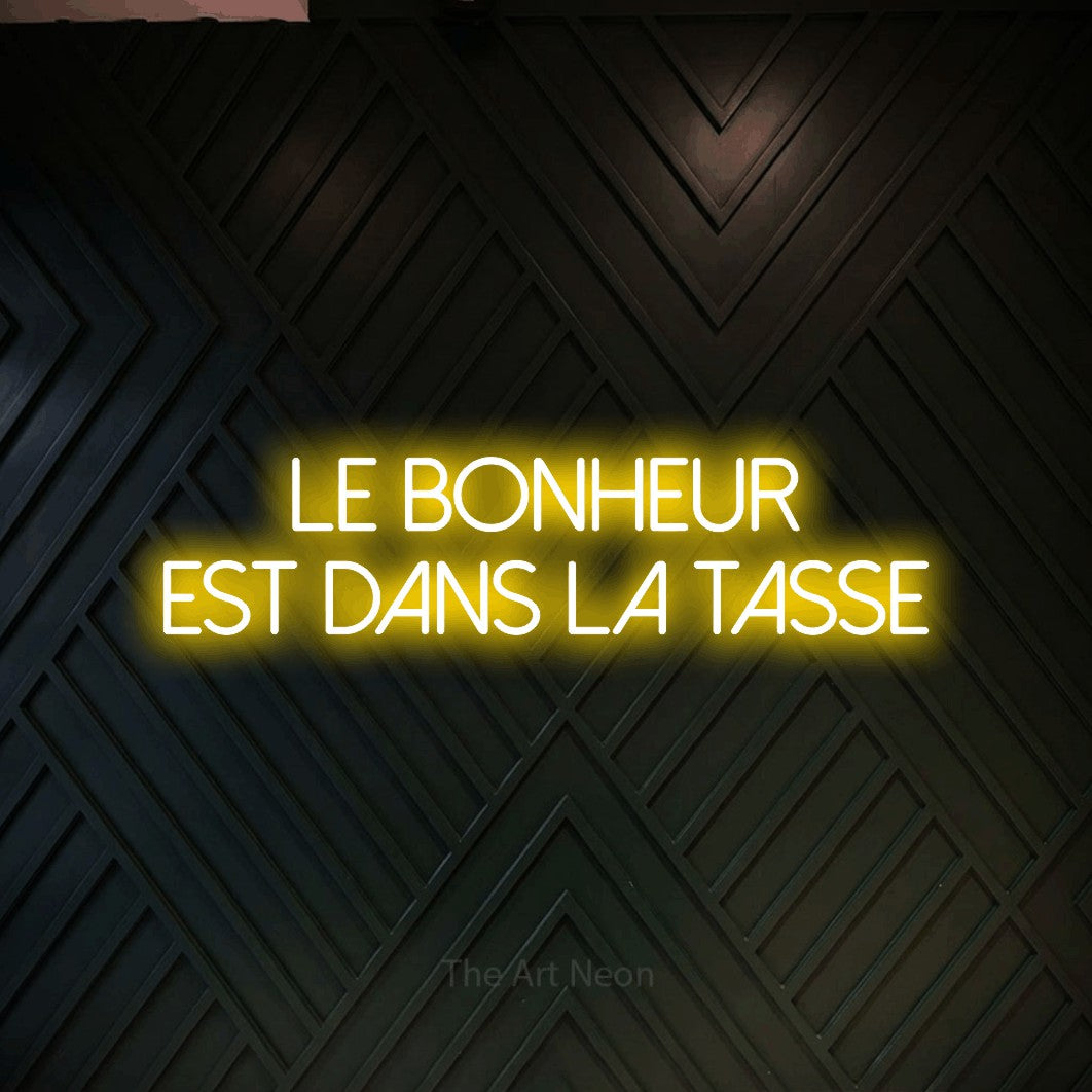 “Le bonheur est dans la tasse” LED neon sign in yellow, with thin sans-serif lettering across 2 lines, shown against a black chevron-patterned wood wall.