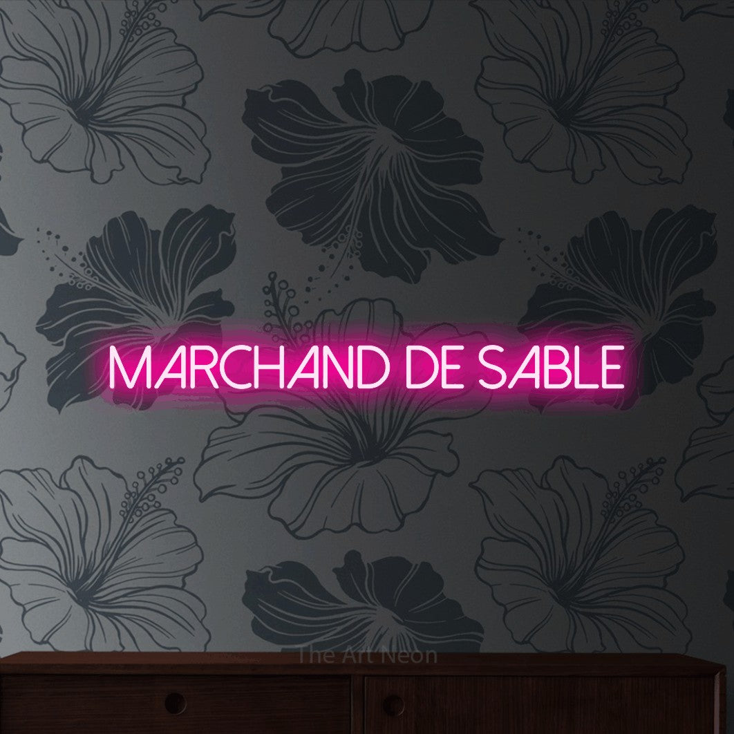 “Marchand de sable” LED neon sign in hot pink, with thin sans-serif lettering, shown against a grey floral-patterned wall.