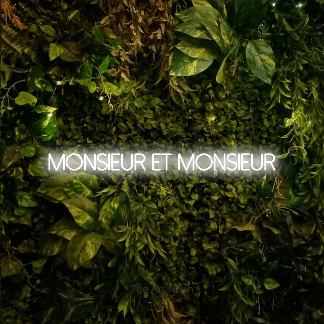 “Monsieur et monsieur” LED neon sign in cool white, with thin sans-serif lettering, shown against bright green and yellow foliage.