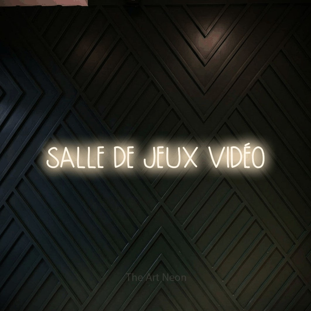 “Salle de jeux vidéo” LED neon sign in warm white, with narrow hand-drawn lettering, shown against a black chevron-patterned wood wall.