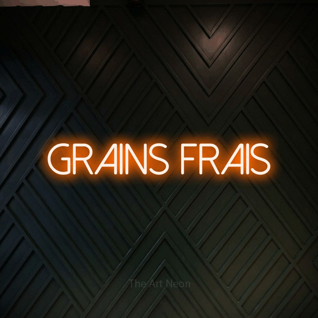 “Grains frais” LED neon sign in orange, with thin sans-serif lettering, shown against a black chevron-patterned wood wall.