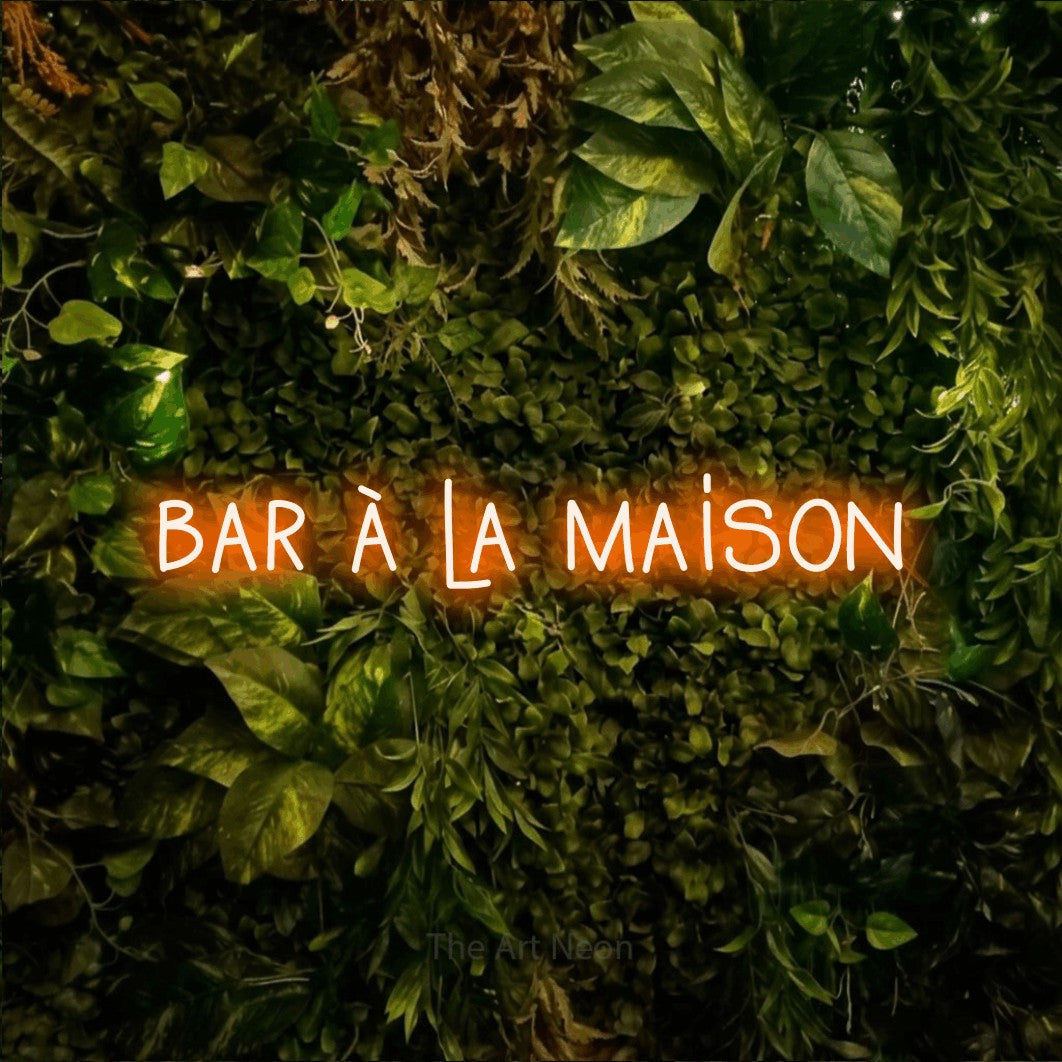 “Bar à la maison” LED neon sign in orange, with narrow hand-drawn lettering, shown against bright green and yellow foliage.