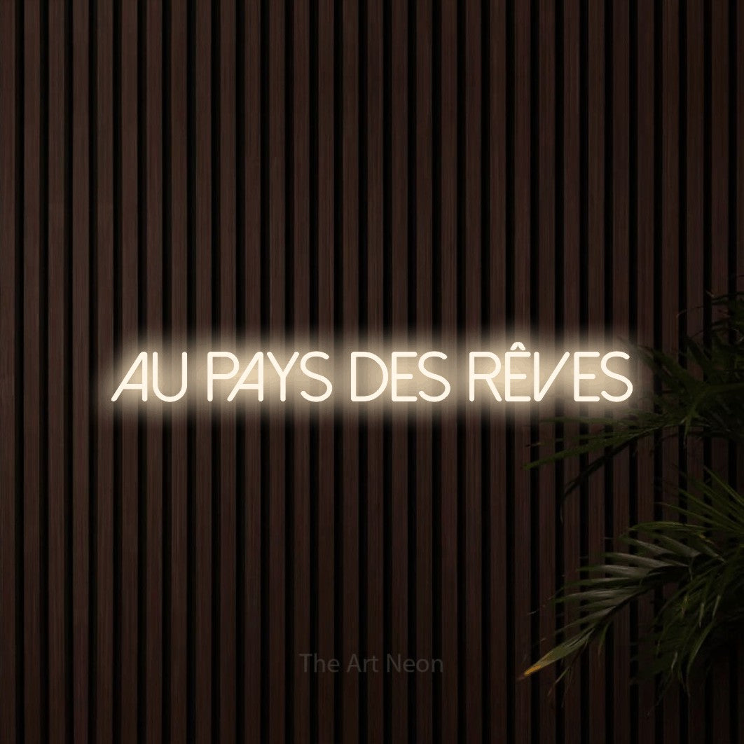 “Au pays des rêves” LED neon sign in warm white, with thin sans-serif lettering, shown against dark wooden wall slats.