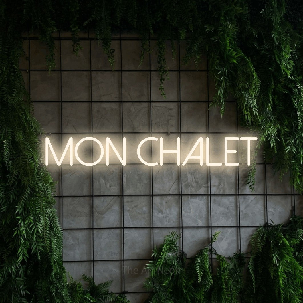 “Mon chalet” LED neon sign in warm white, with thin sans-serif lettering, shown against an ivy-covered concrete grid.