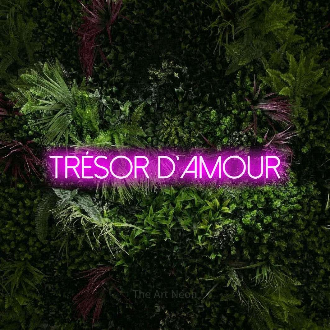 “Trésor d'amour” LED neon sign in pink, with thin sans-serif lettering, shown against a plant wall with pink flowers.
