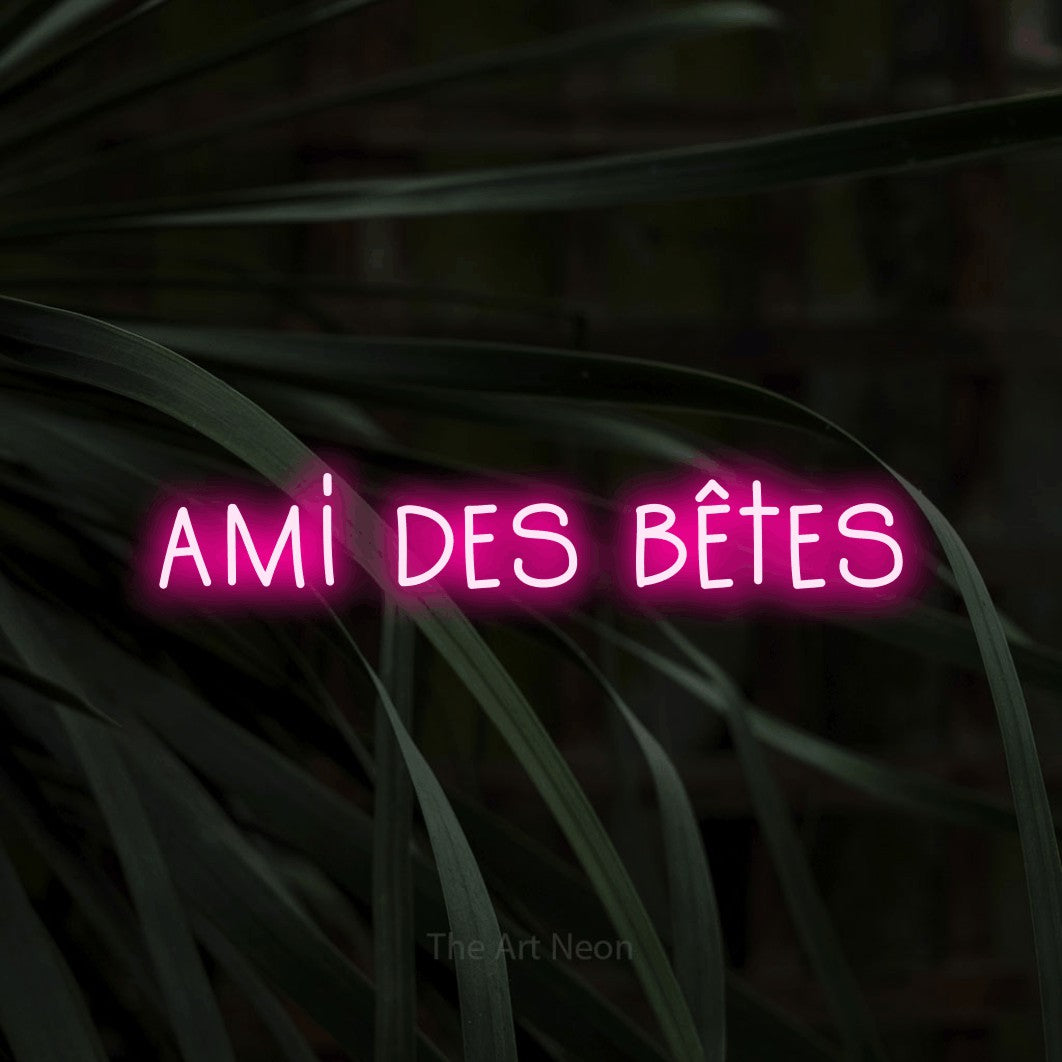 “Ami des bêtes” LED neon sign in hot pink, with narrow hand-drawn lettering, shown against a close-up of dark foliage.