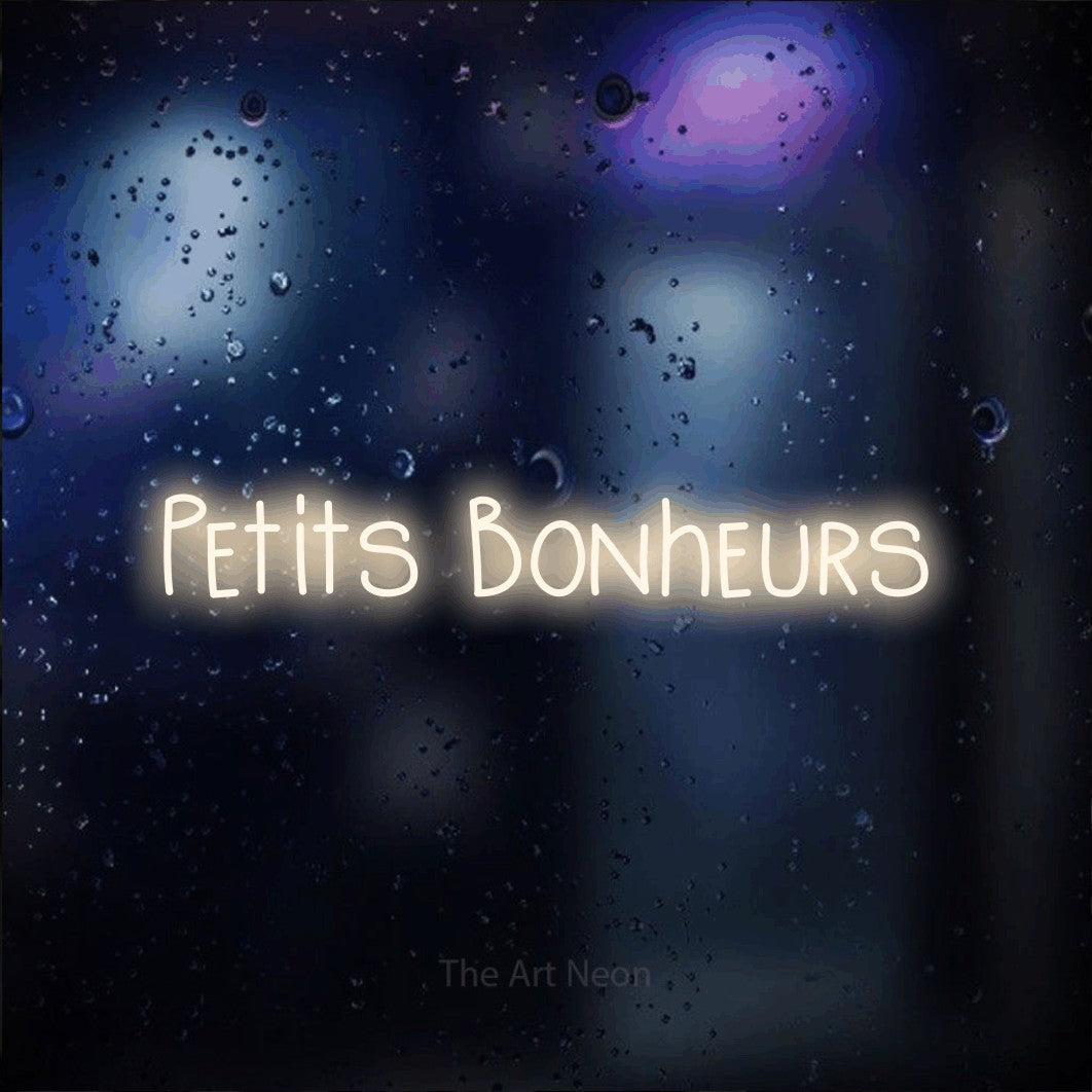 “Petits bonheurs” LED neon sign in warm white, with narrow hand-drawn lettering, shown against glass with blue and purple reflections.