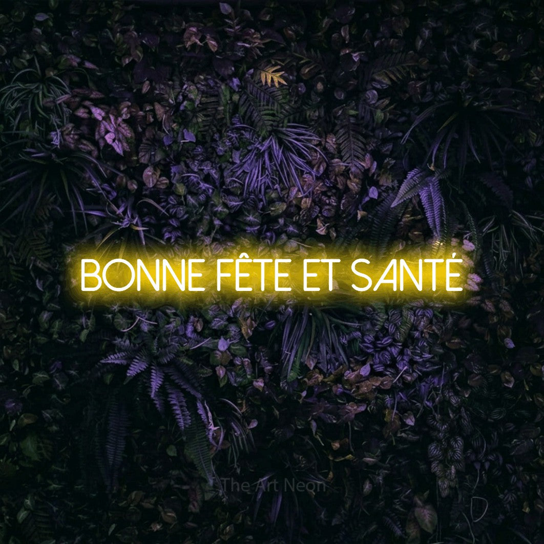 “Bonne fête et santé” LED neon sign in yellow, with thin sans-serif lettering, shown against a dark wall with purple flowers.