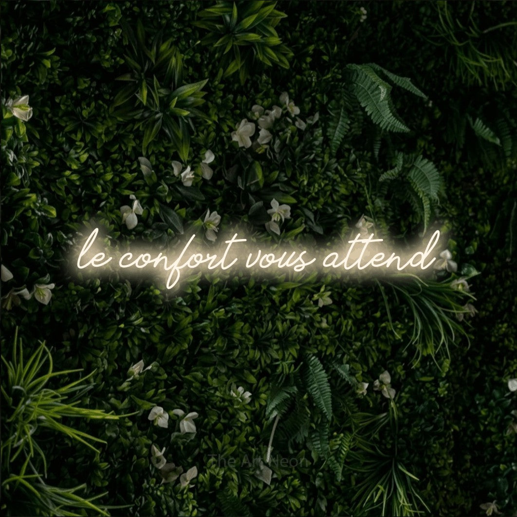 “Le confort vous attend” LED neon sign in warm white, with cursive lettering, shown against a dense green plant wall.