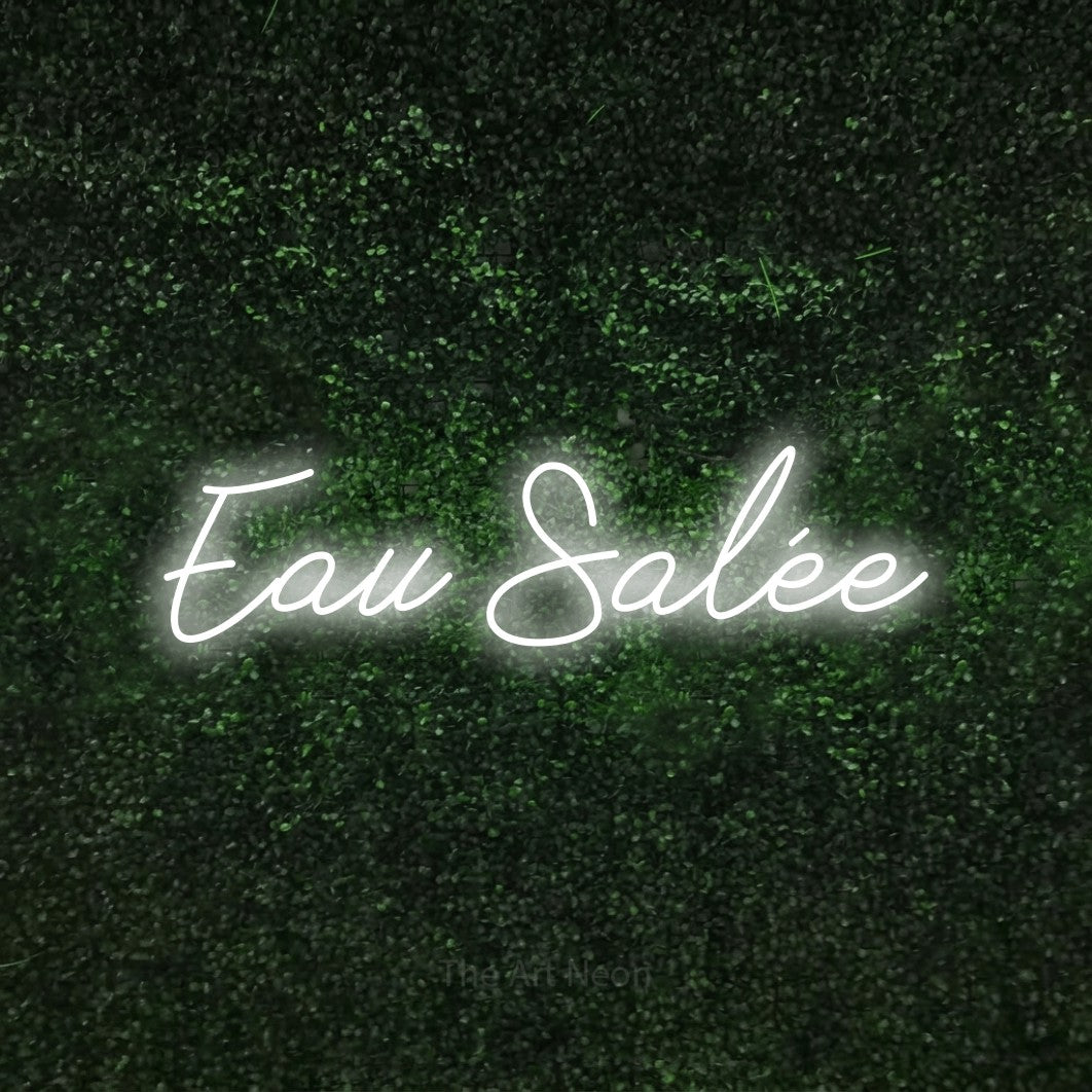 “Eau salée” LED neon sign in cool white, with cursive lettering, shown against a textured green hedge.