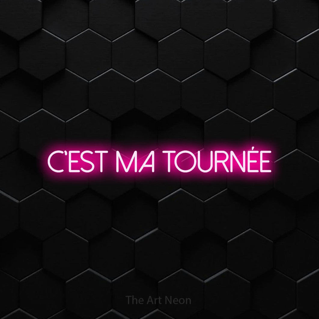 “C'est ma tournée” LED neon sign in hot pink, with thin sans-serif lettering, shown against black hexagonal wall tiles.