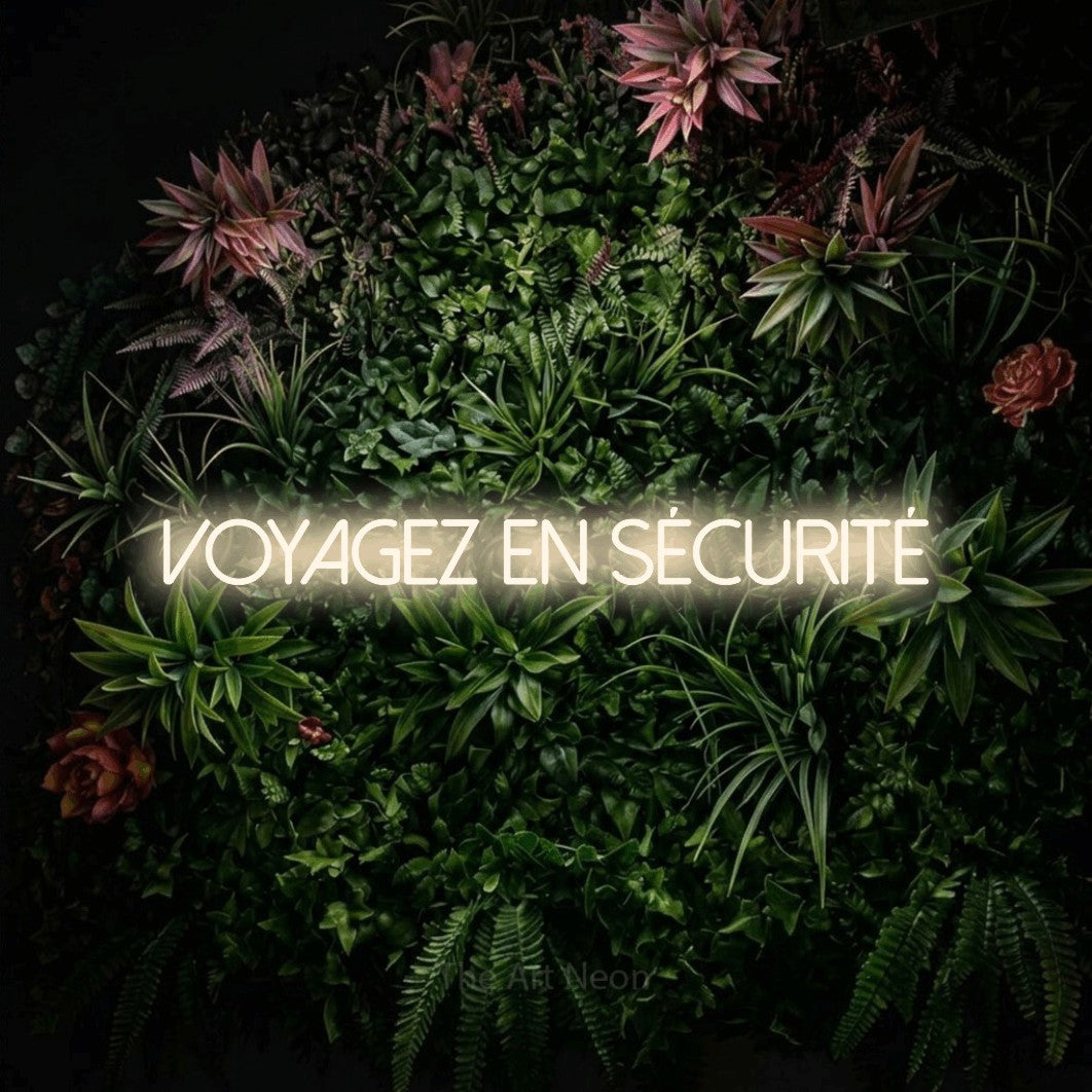 “Voyagez en sécurité” LED neon sign in warm white, with thin sans-serif lettering, shown against a flowering plant wall.