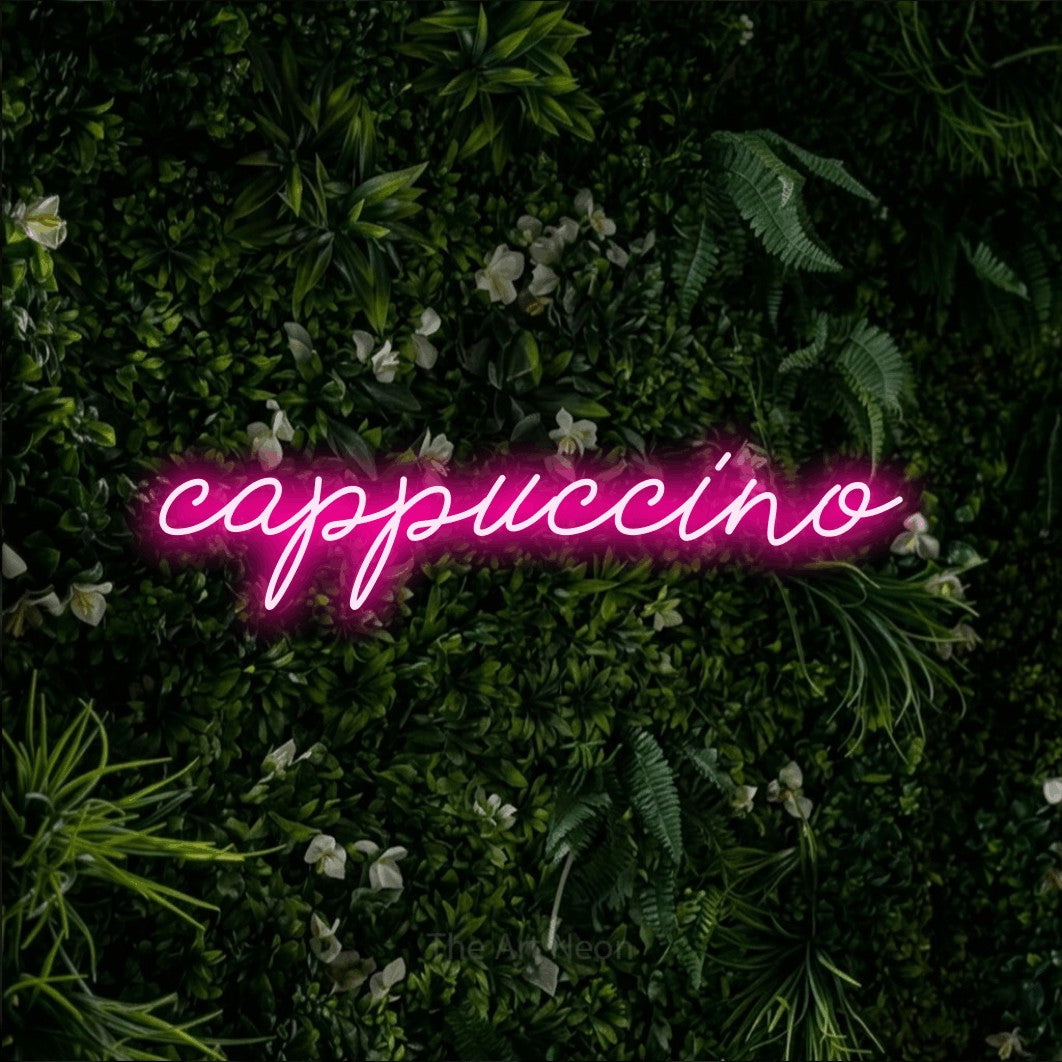 “Cappuccino” LED neon sign in hot pink, with cursive lettering, shown against a dense green plant wall.