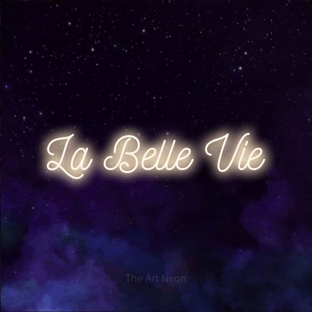 “La belle vie” LED neon sign in warm white, with ornate cursive lettering, shown against a purple galaxy backdrop.
