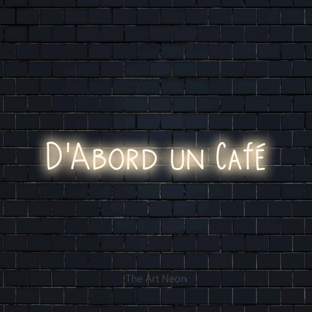 “D'abord un café” LED neon sign in warm white, with narrow hand-drawn lettering, shown against a dark navy brick wall.