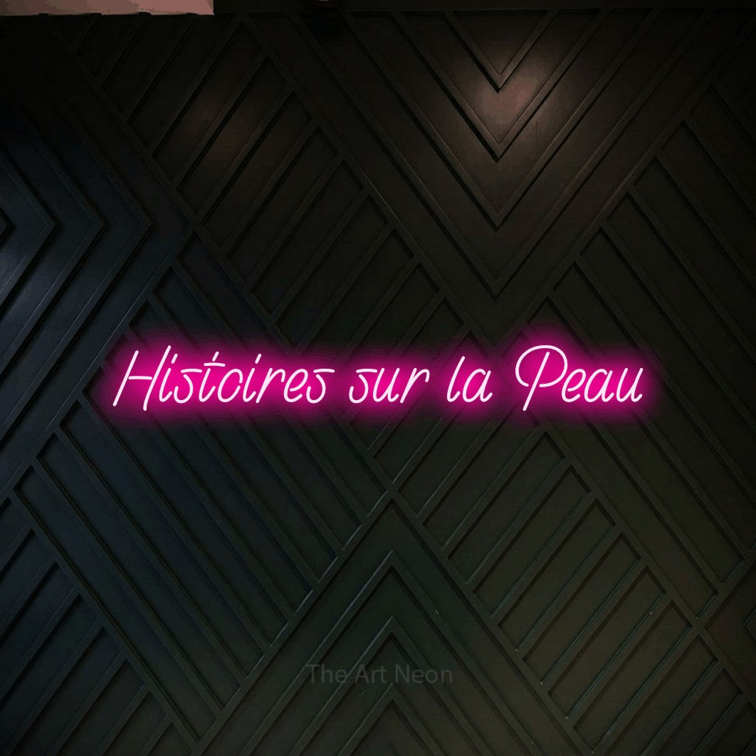 “Histoires sur la peau” LED neon sign in hot pink, with slanted handwritten lettering, shown against a black chevron-patterned wood wall.