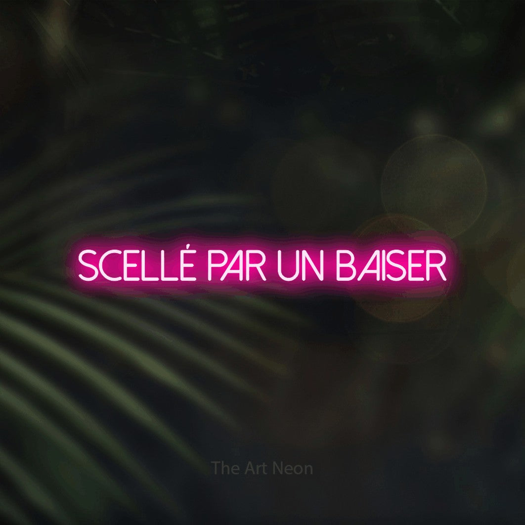 “Scellé par un baiser” LED neon sign in hot pink, with thin sans-serif lettering, shown against a blurred palm and bokeh-light backdrop.