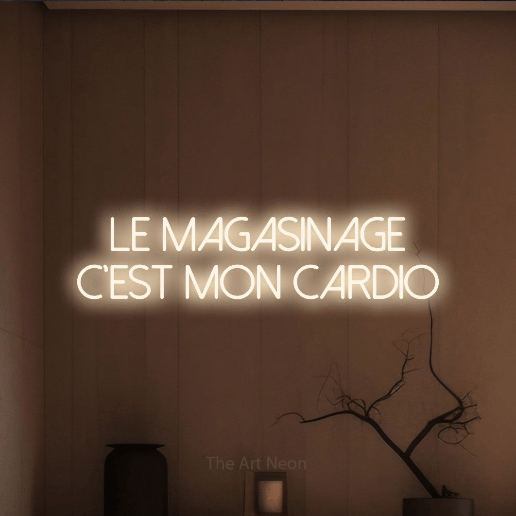 “Le magasinage c'est mon cardio” LED neon sign in warm white, with thin sans-serif lettering across 2 lines, shown against a simple earth-toned interior.