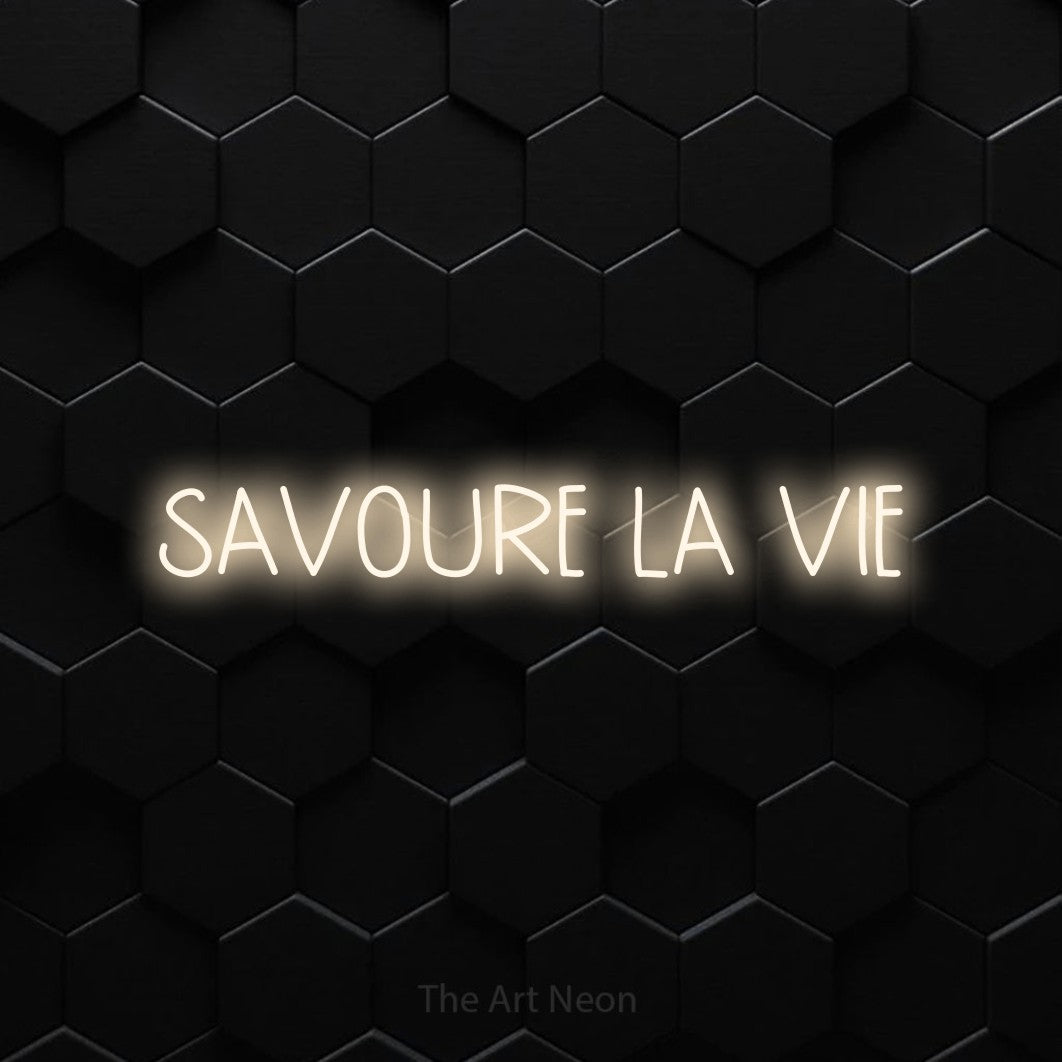 “Savoure la vie” LED neon sign in warm white, with narrow hand-drawn lettering, shown against black hexagonal wall tiles.