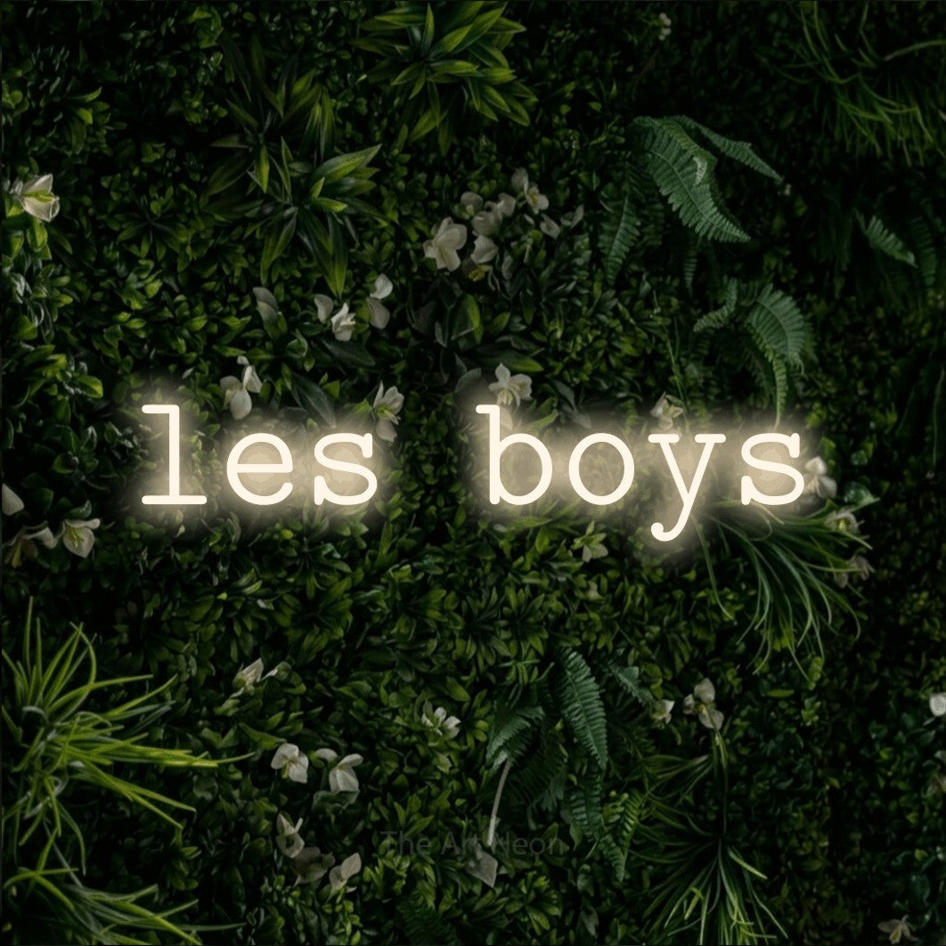 “Les boys” LED neon sign in warm white, with typewriter-style lettering, shown against a dense green plant wall.