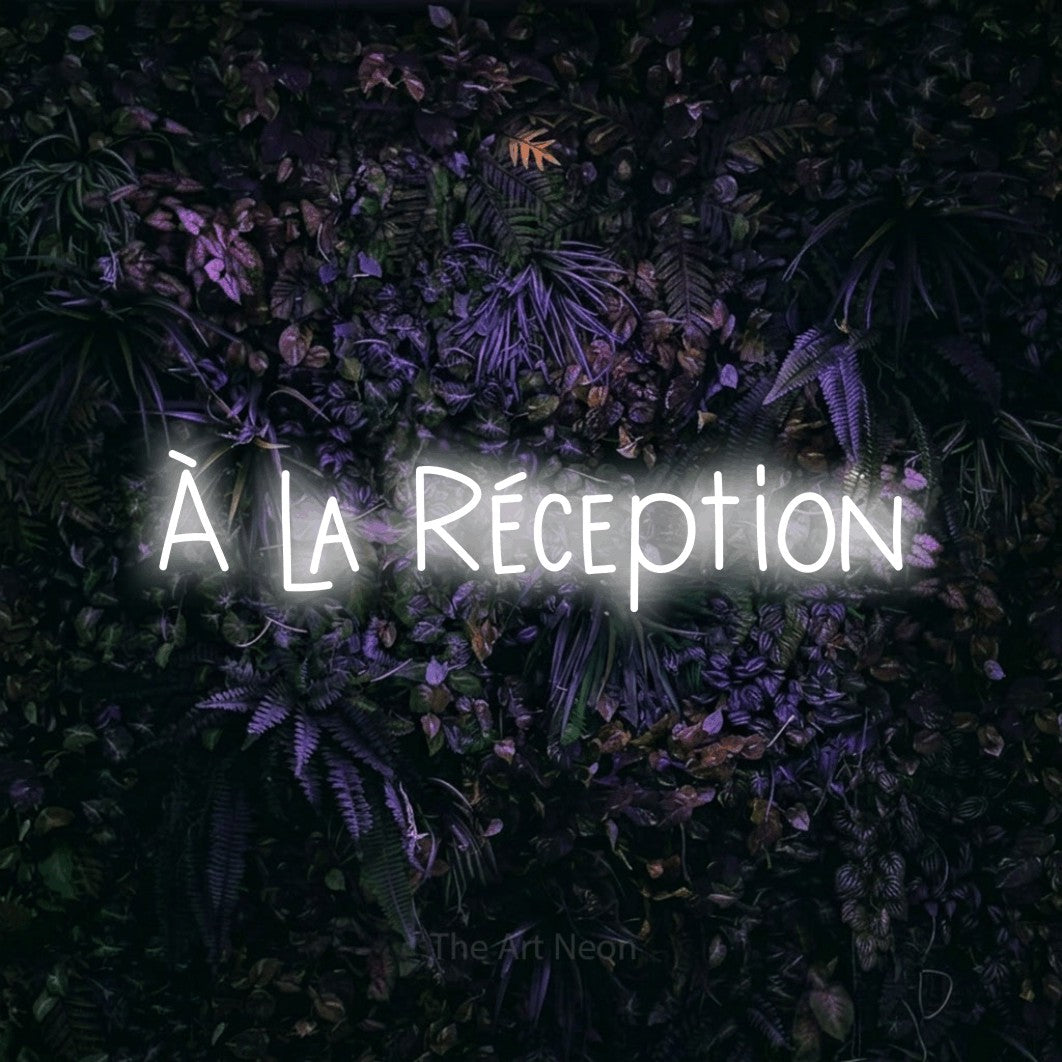 “À la réception” LED neon sign in cool white, with narrow hand-drawn lettering, shown against a dark wall with purple flowers.