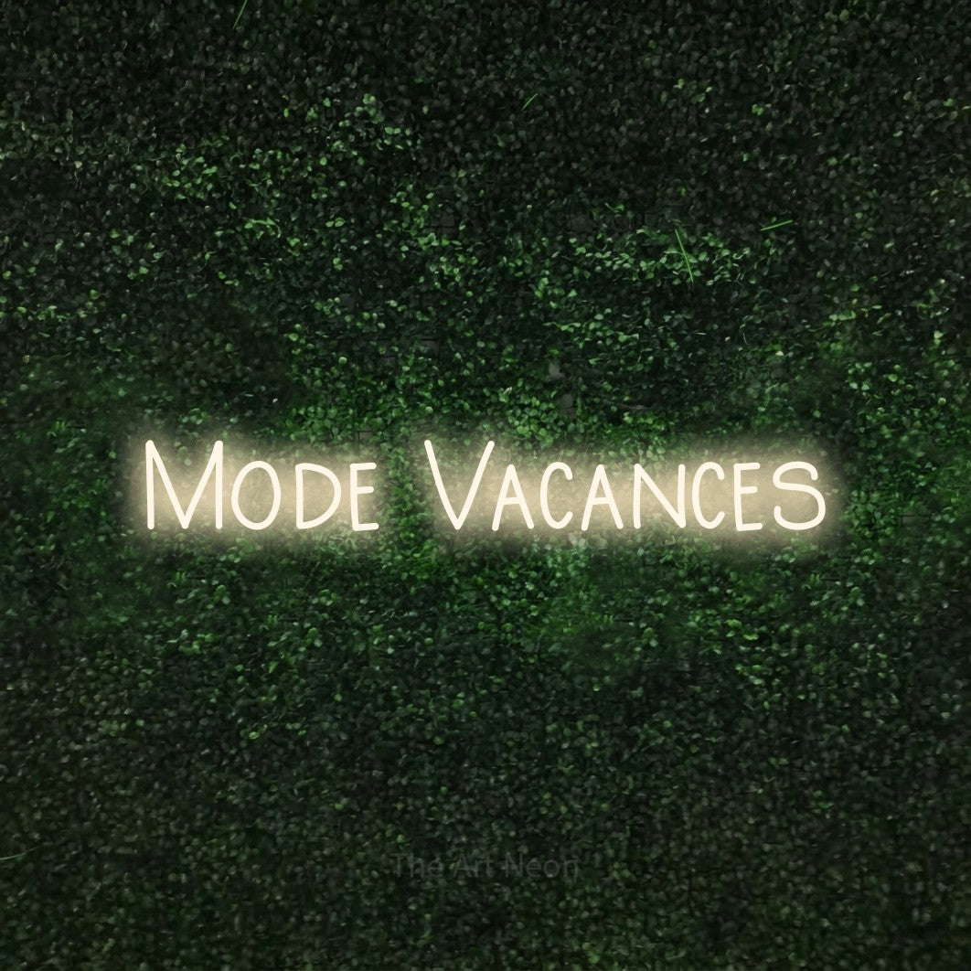 “Mode vacances” LED neon sign in warm white, with narrow hand-drawn lettering, shown against a textured green hedge.