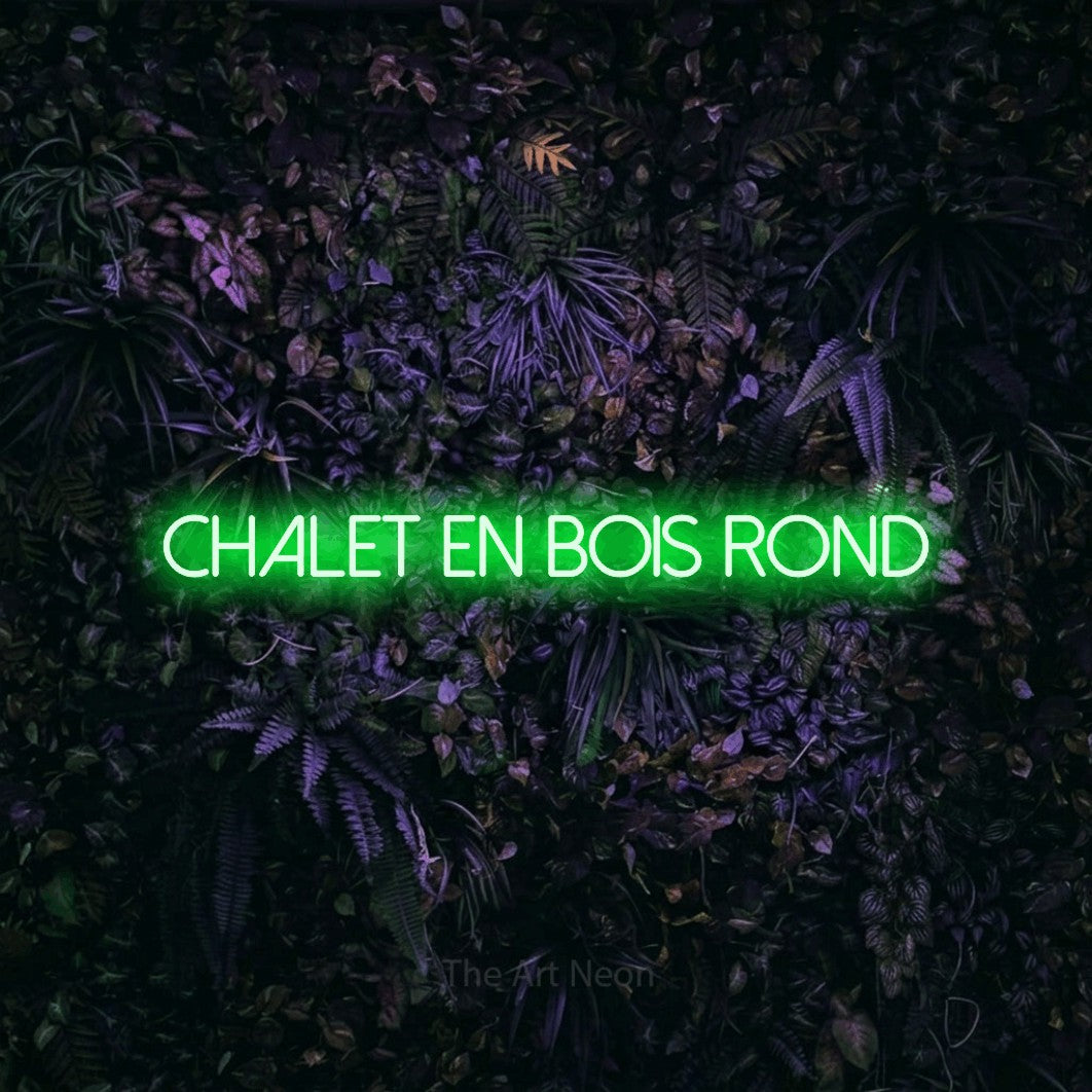 “Chalet en bois rond” LED neon sign in green, with thin sans-serif lettering, shown against a dark wall with purple flowers.