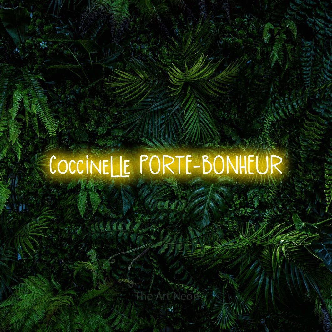 “Coccinelle porte-bonheur” LED neon sign in yellow, with narrow hand-drawn lettering, shown against dense tropical foliage.