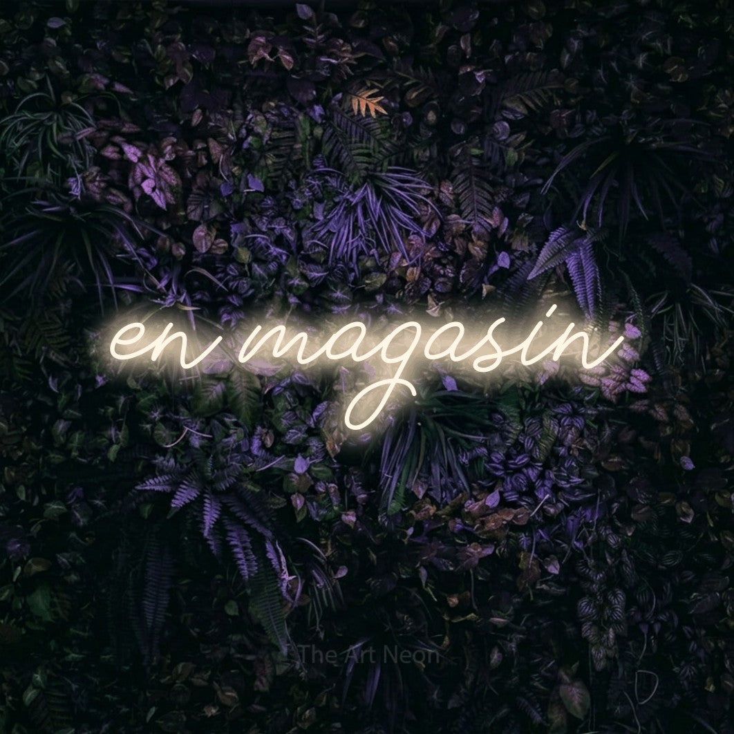 “En magasin” LED neon sign in warm white, with cursive lettering, shown against a dark wall with purple flowers.