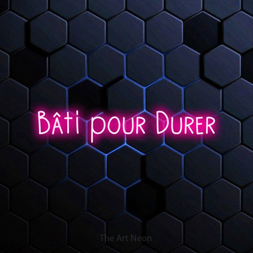“Bâti pour durer” LED neon sign in hot pink, with narrow hand-drawn lettering, shown against a dark blue honeycomb pattern.