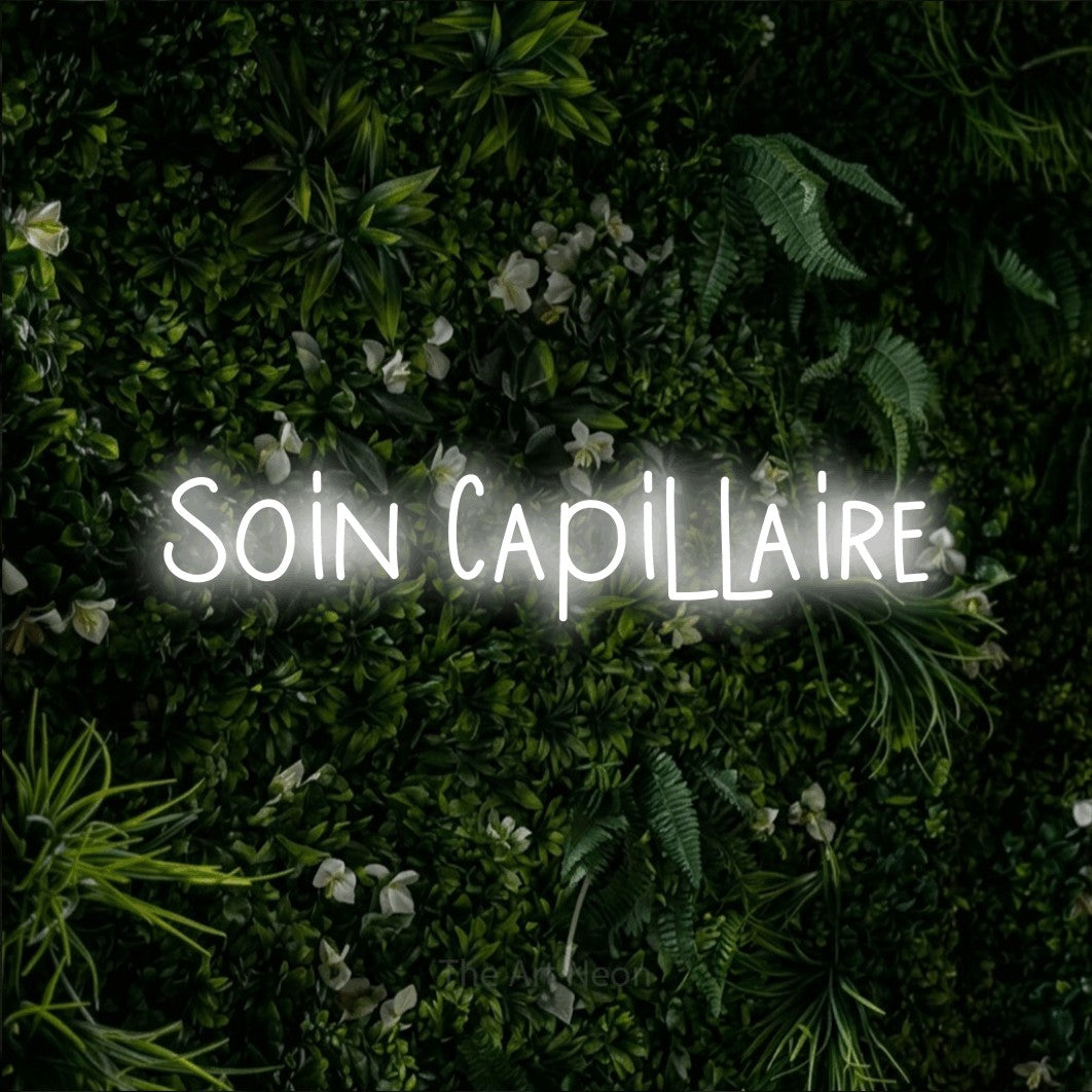 “Soin capillaire” LED neon sign in cool white, with narrow hand-drawn lettering, shown against a dense green plant wall.