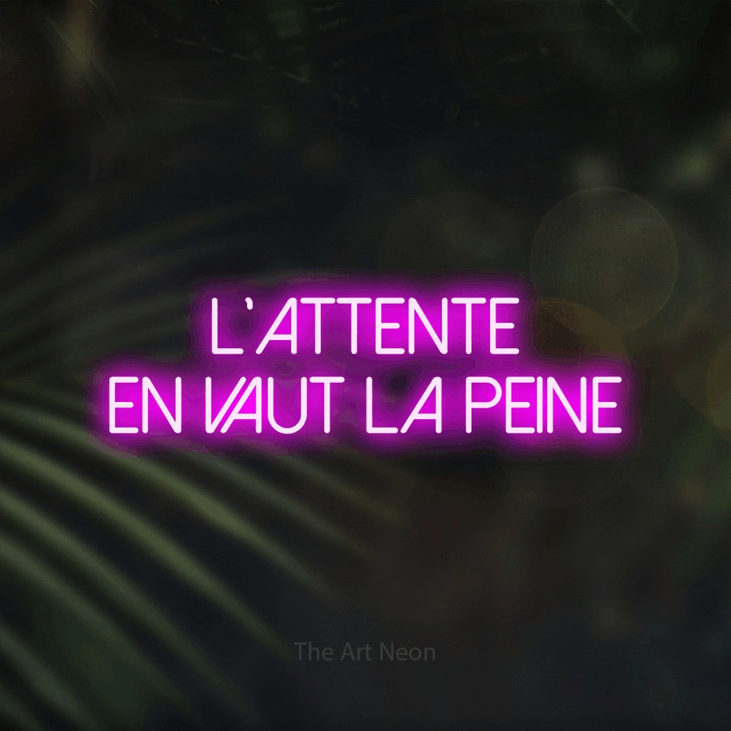 “L'attente en vaut la peine” LED neon sign in pink, with thin sans-serif lettering across 2 lines, shown against a blurred palm and bokeh-light backdrop.