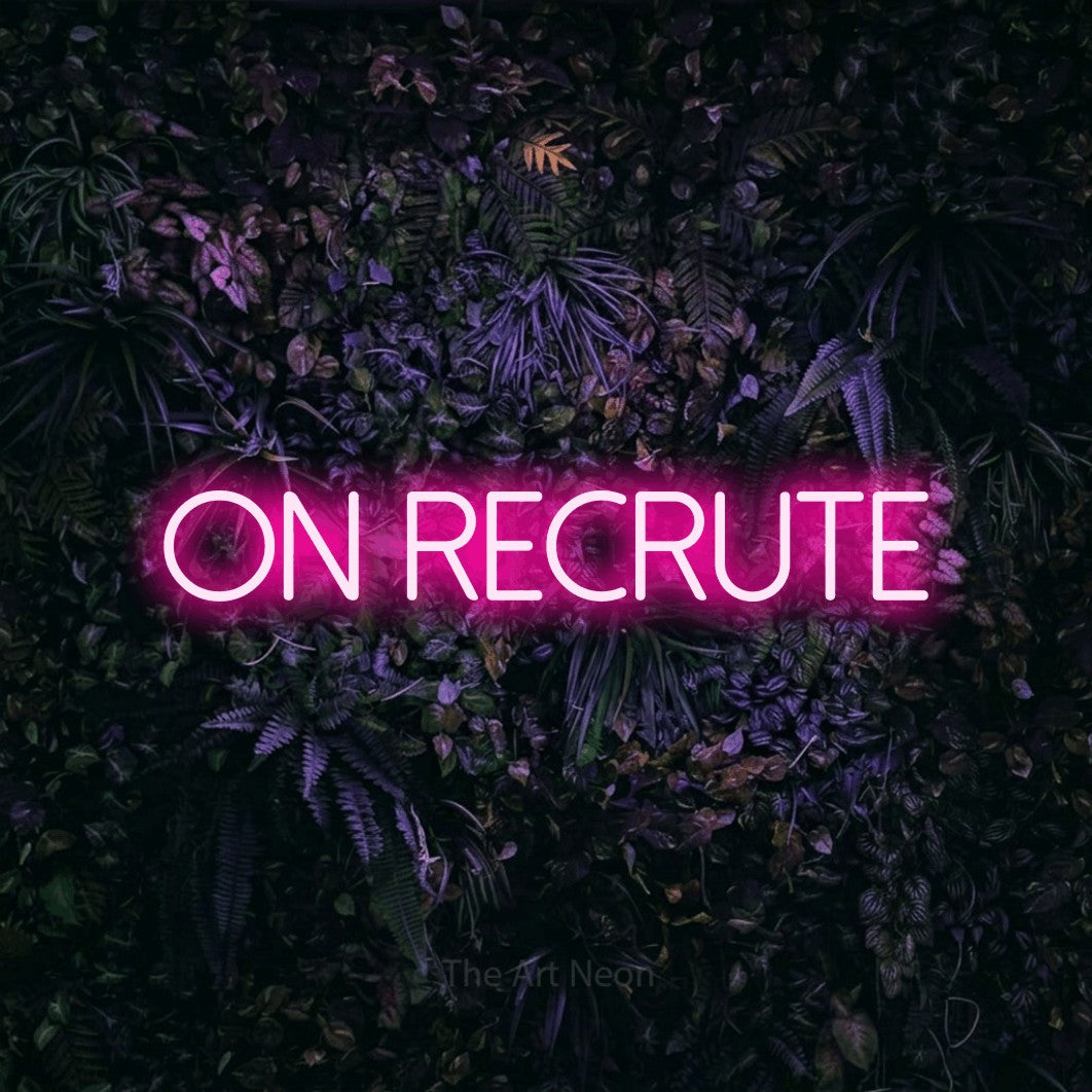 “On recrute” LED neon sign in hot pink, with thin sans-serif lettering, shown against a dark wall with purple flowers.