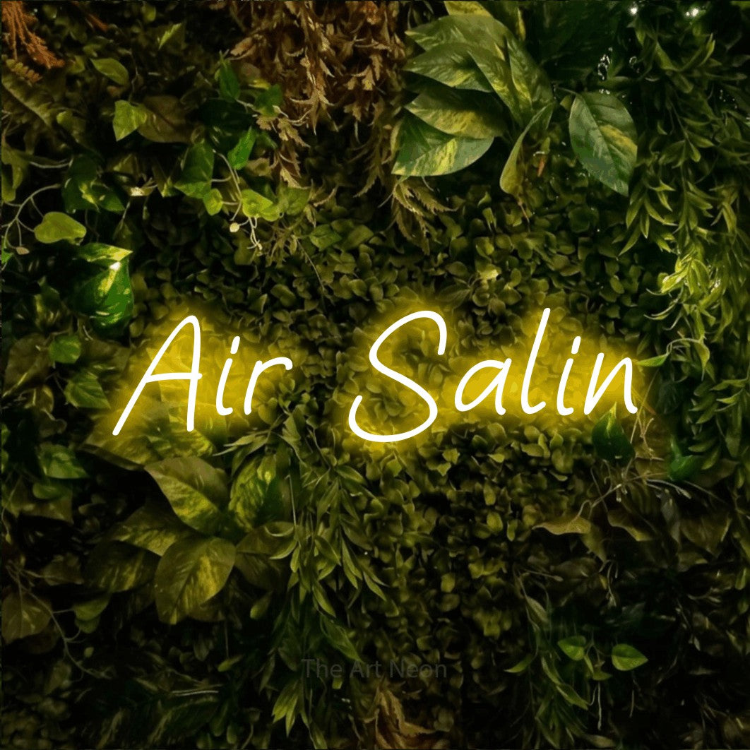 “Air salin” LED neon sign in yellow, with slanted handwritten lettering, shown against bright green and yellow foliage.