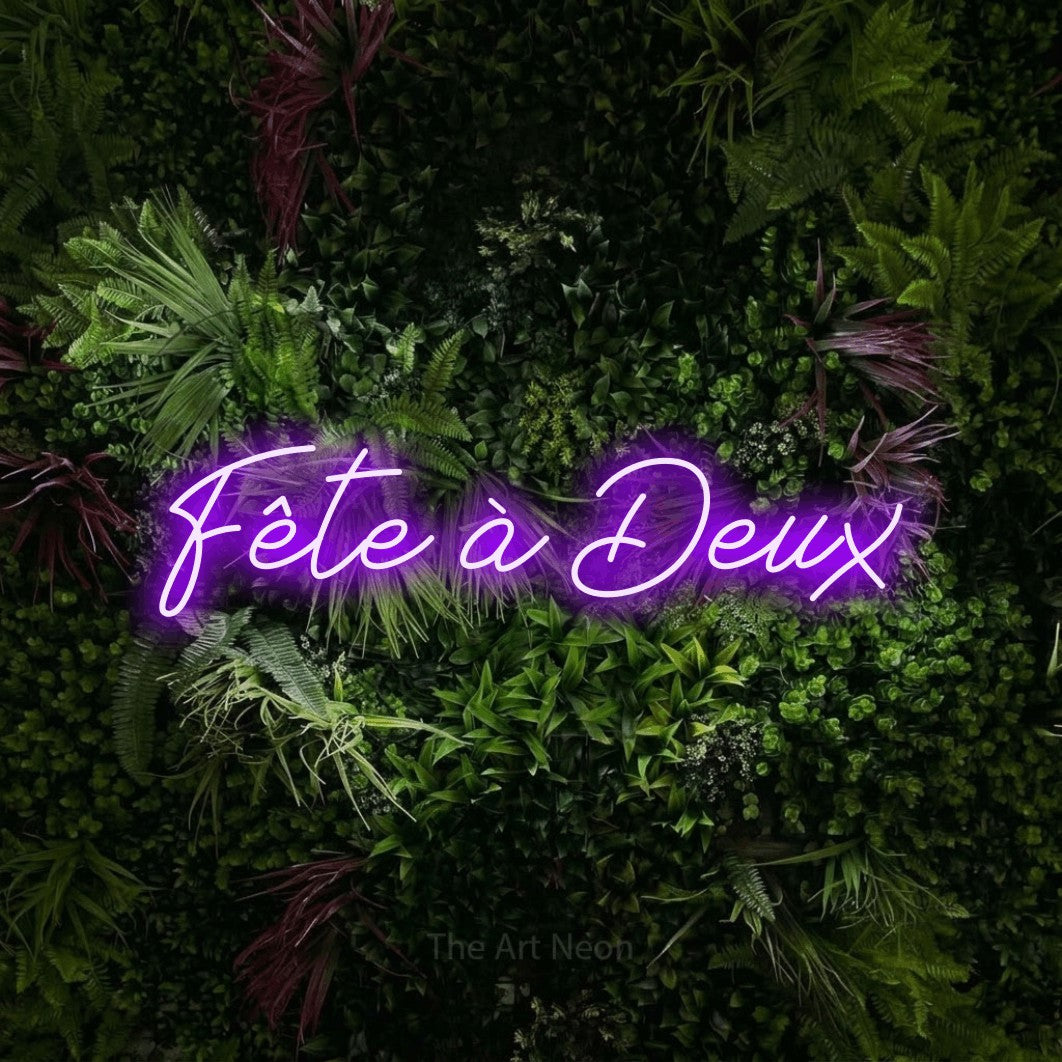 “Fête à deux” LED neon sign in purple, with cursive lettering, shown against a plant wall with pink flowers.