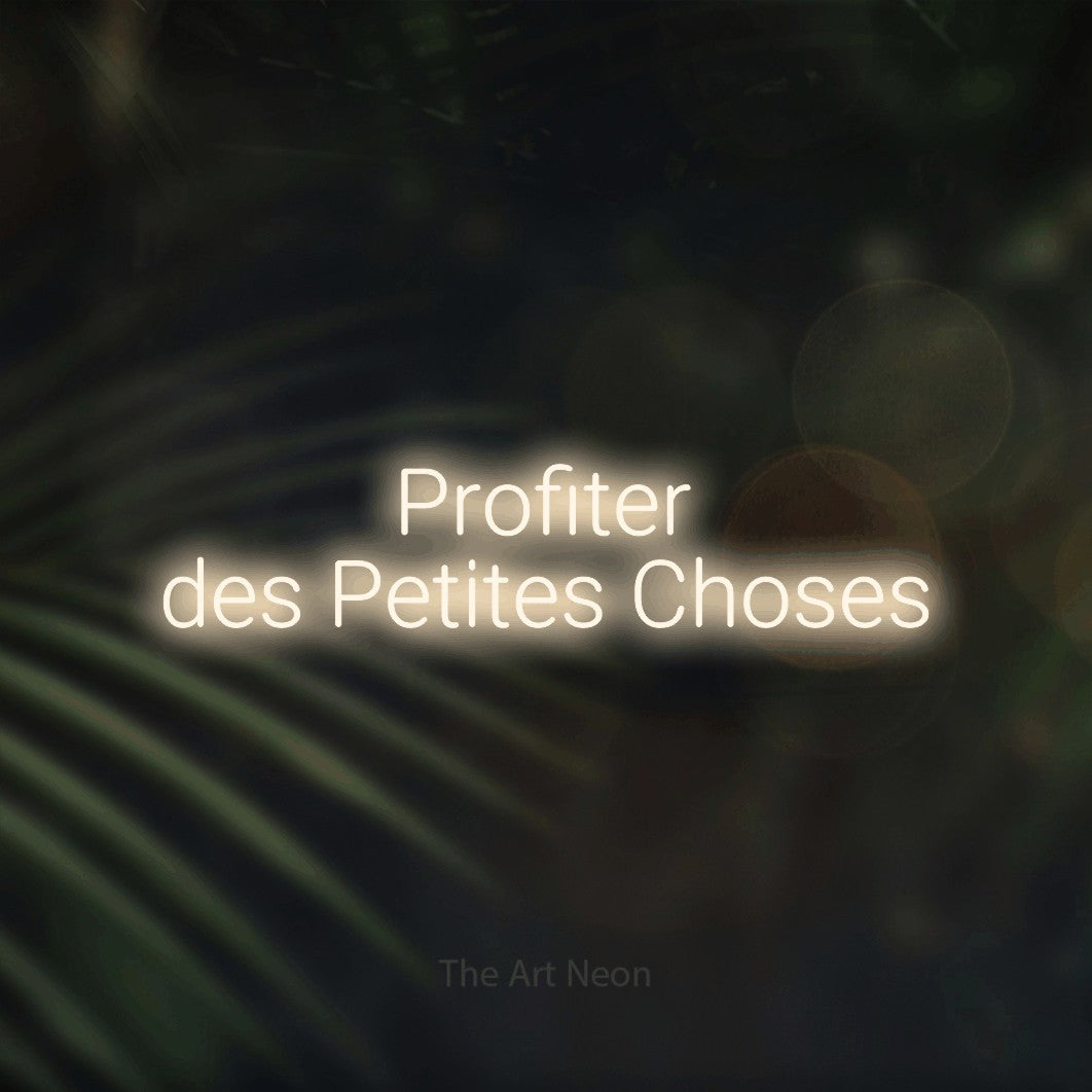 “Profiter des petites choses” LED neon sign in warm white, with rounded sans-serif lettering across 2 lines, shown against a blurred palm and bokeh-light backdrop.