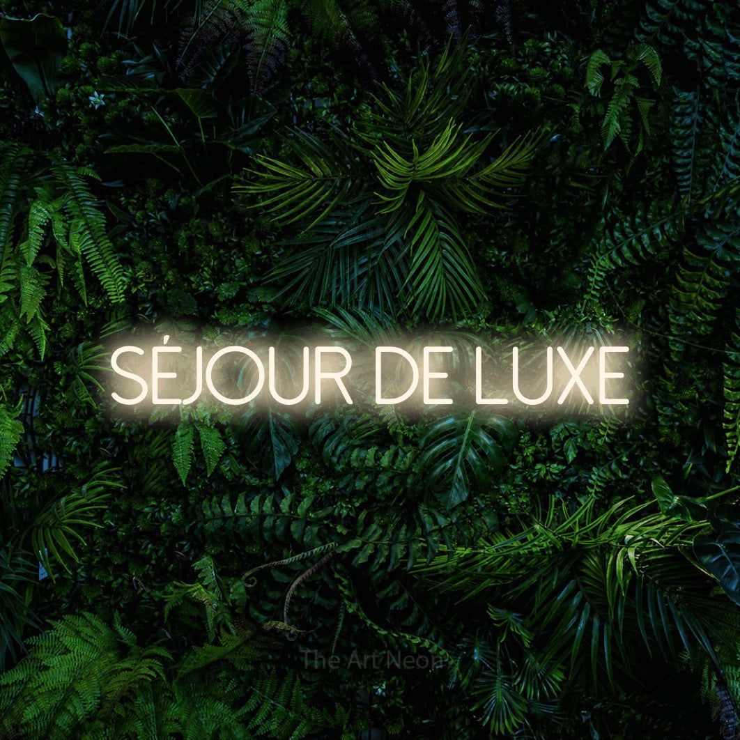 “Séjour de luxe” LED neon sign in warm white, with thin sans-serif lettering, shown against dense tropical foliage.