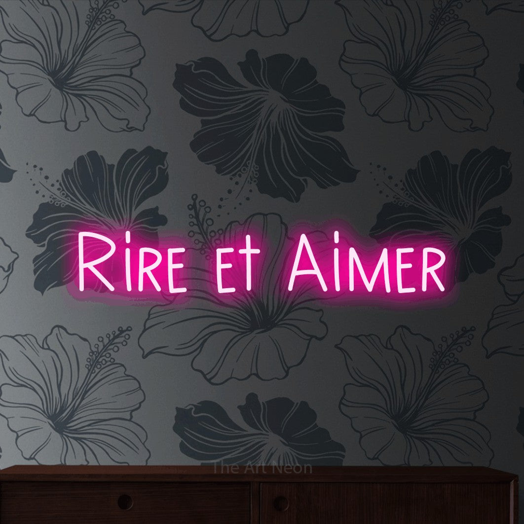 “Rire et aimer” LED neon sign in hot pink, with narrow hand-drawn lettering, shown against a grey floral-patterned wall.