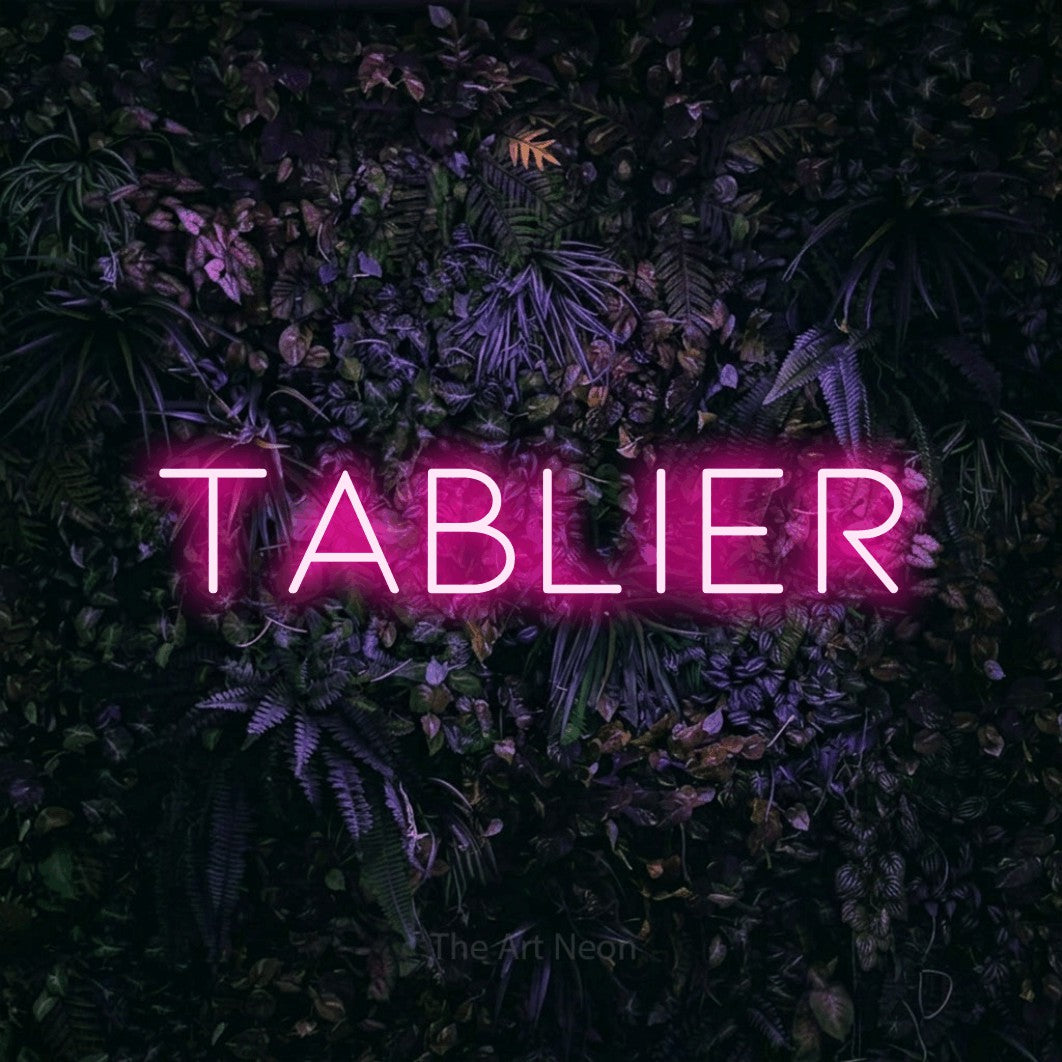 “Tablier” LED neon sign in hot pink, with thin, widely spaced lettering, shown against a dark wall with purple flowers.