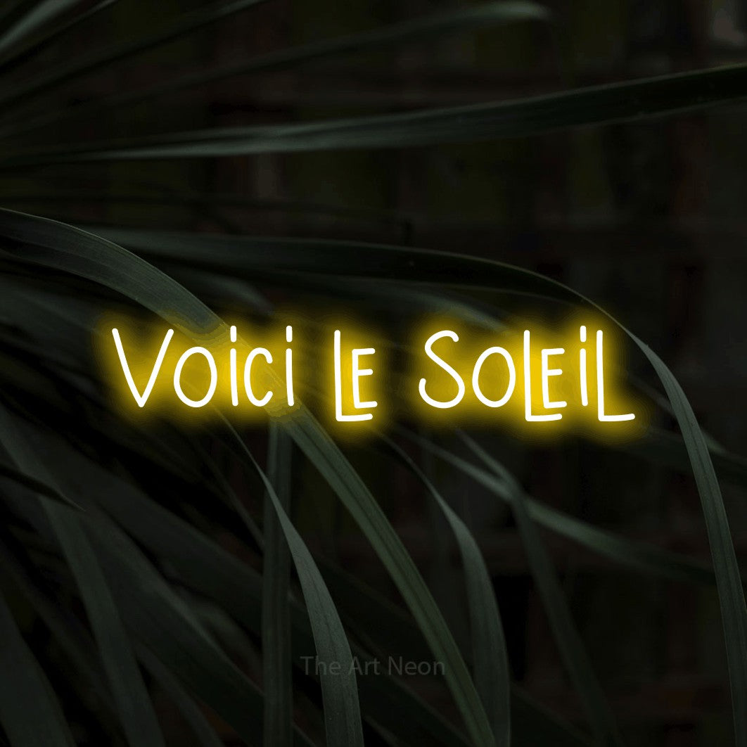 “Voici le soleil” LED neon sign in yellow, with narrow hand-drawn lettering, shown against a close-up of dark foliage.