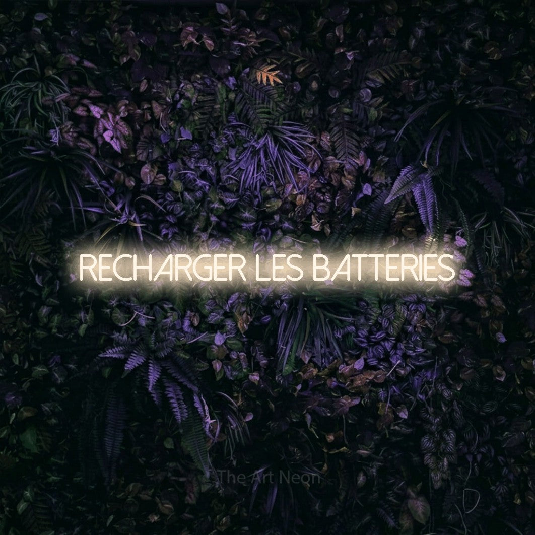 “Recharger les batteries” LED neon sign in warm white, with thin sans-serif lettering, shown against a dark wall with purple flowers.