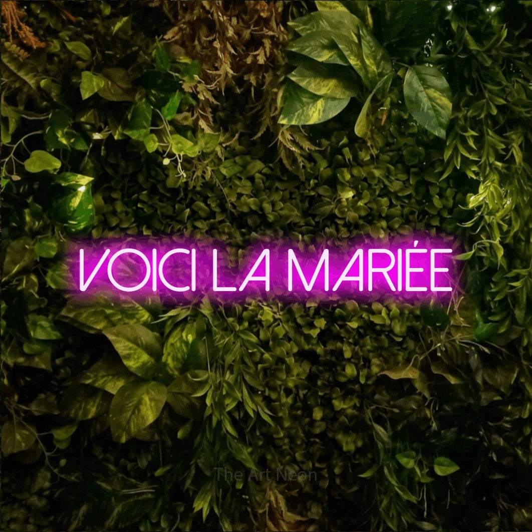 “Voici la mariée” LED neon sign in pink, with thin sans-serif lettering, shown against bright green and yellow foliage.