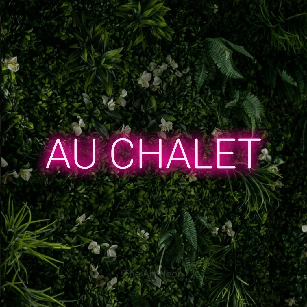 Composition en néon LED « Au chalet » en rose vif, en lettres sans empattement arrondies, mise en valeur sur un mur végétal vert et dense, pour un décor canadien.