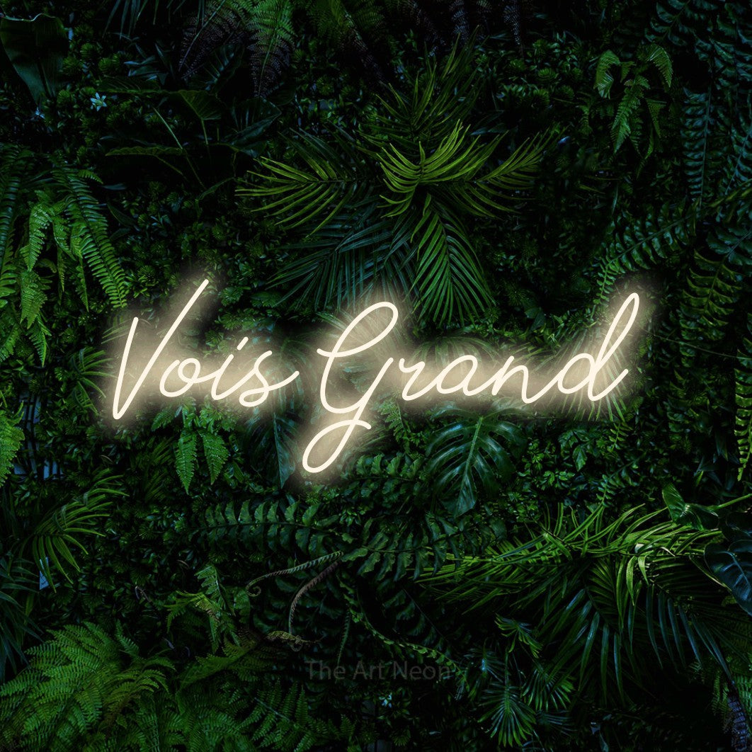 “Vois grand” LED neon sign in warm white, with cursive lettering, shown against dense tropical foliage.