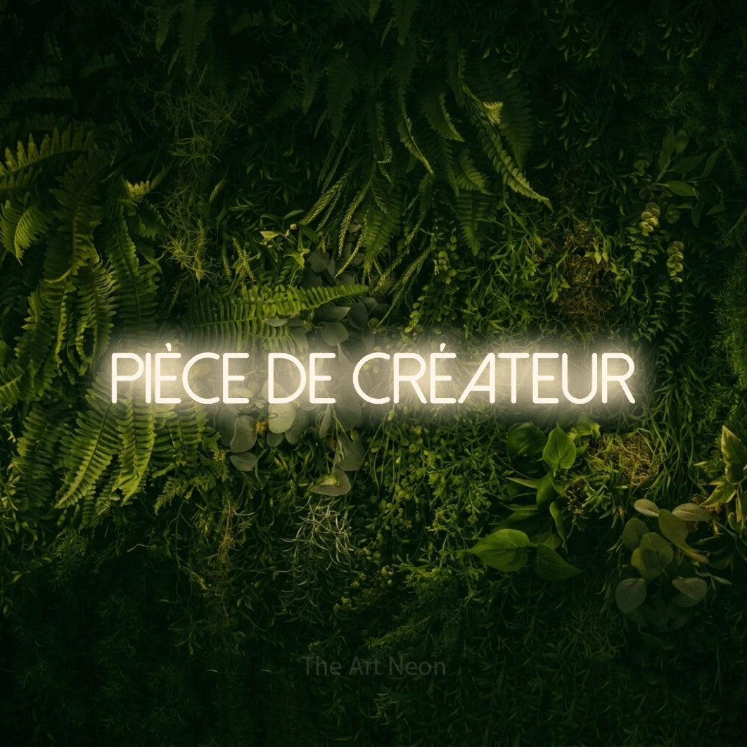 “Pièce de créateur” LED neon sign in warm white, with thin sans-serif lettering, shown against a dark moss and forest backdrop.