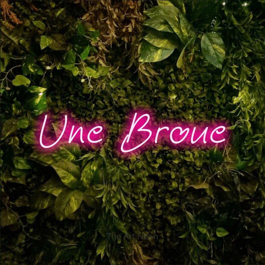 “Une broue” LED neon sign in hot pink, with slanted handwritten lettering, shown against bright green and yellow foliage.