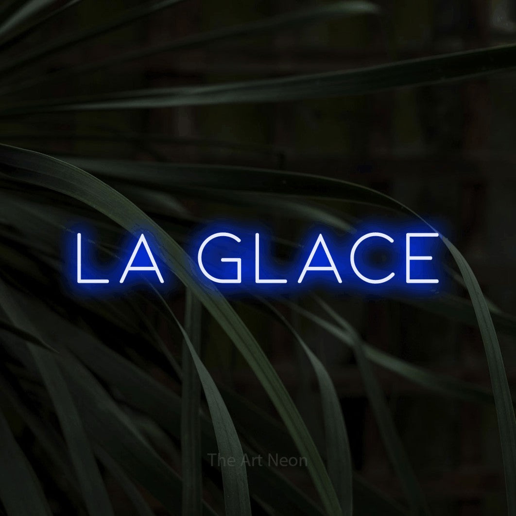 “La glace” LED neon sign in blue, with thin, widely spaced lettering, shown against a close-up of dark foliage.
